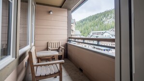 Ein Balkon mit einer Holzbänkl und einem Tisch, Blick auf einen bewaldeten Berg und Gebäude im Hintergrund.