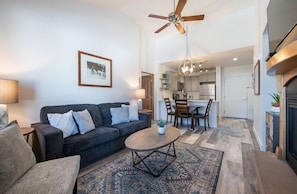 A living room with a sofa, coffee table, and a ceiling fan. There is a kitchen visible in the background.