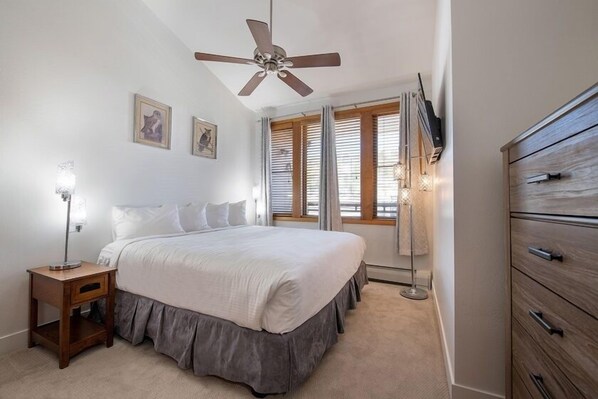 Un dormitorio con cama, mesita de noche, cómoda, ventilador de techo y ventana con persianas.