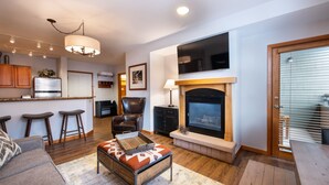 A modern living room with a fireplace, a kitchen area with bar stools, and a seating area with a sofa and ottoman. - Zephyr Mountain Lodge, Condo | 2 Bedroom (Select-Rated Condo 1415) (Winter Park)