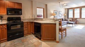 A kitchen with wooden cabinets, a black stove, and a microwave. There is a dining area with a sofa and a table. - Zephyr Mountain Lodge, Condo | Ski Slope View (Select-Rated Condo 2405) (Winter Park)