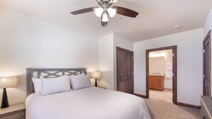 Une chambre à coucher avec un lit, des tables de chevet, un ventilateur de plafond et une salle de bain visible à travers une porte ouverte.