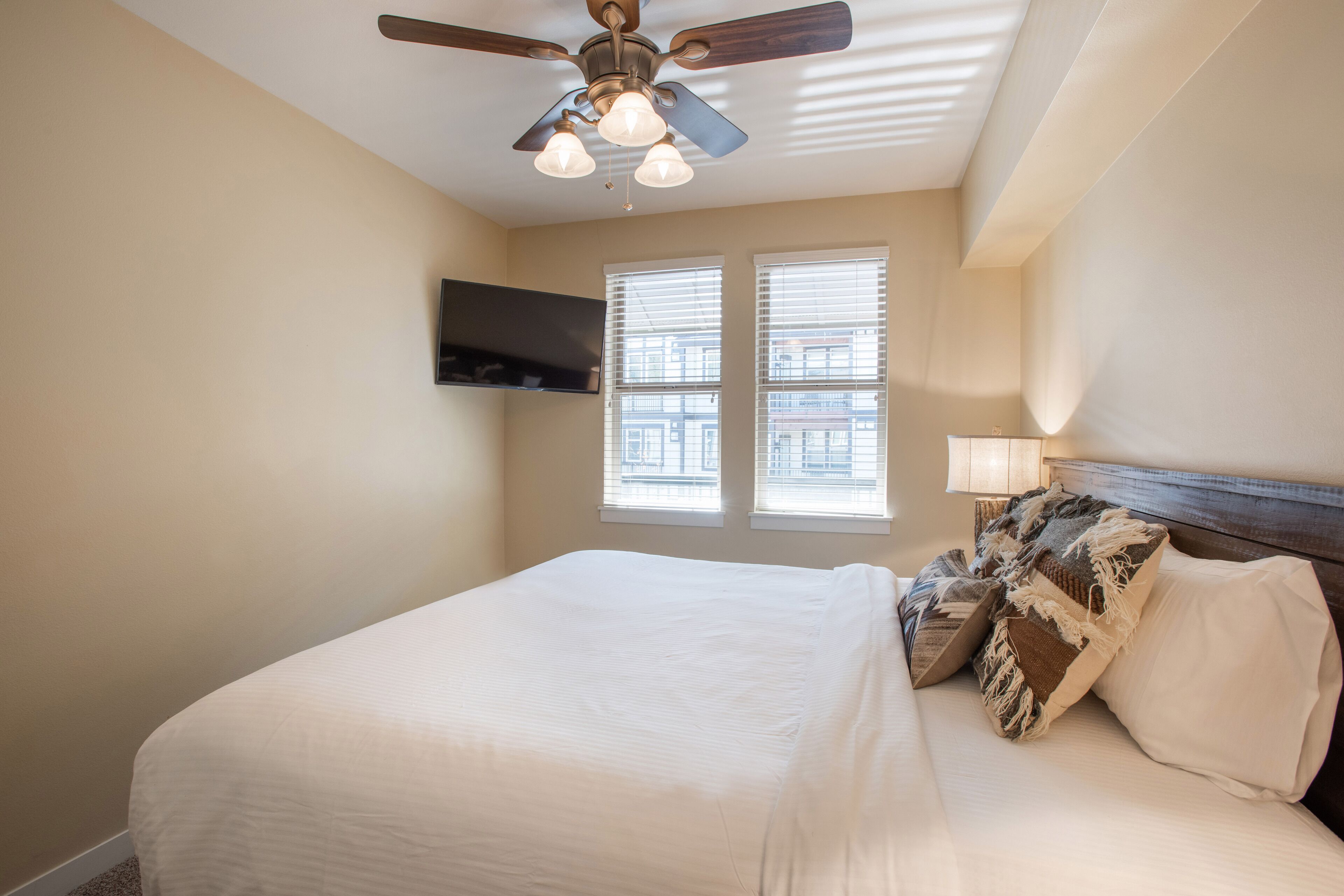 A bedroom with a bed, a ceiling fan, a TV mounted on the wall, and windows with blinds.