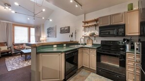 A compact kitchen with a dishwasher, oven, microwave, and a sink. There is a window with curtains, a chair, and a small table. - Zephyr Mountain Lodge, Condo | Ski Storage (Select-Rated Condo 2700) (Winter Park)