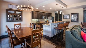 A modern kitchen with dark wooden cabinets, a central island, and a dining area with wooden chairs. - Village View, Townhouse | Fireplace at Parry Peak Lofts (Premium-Rated Condo 106) (Winter Park)