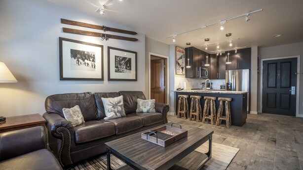Una sala moderna con un sofá de cuero, una mesa de centro de madera y una cocina con armarios oscuros y un refrigerador de acero inoxidable.