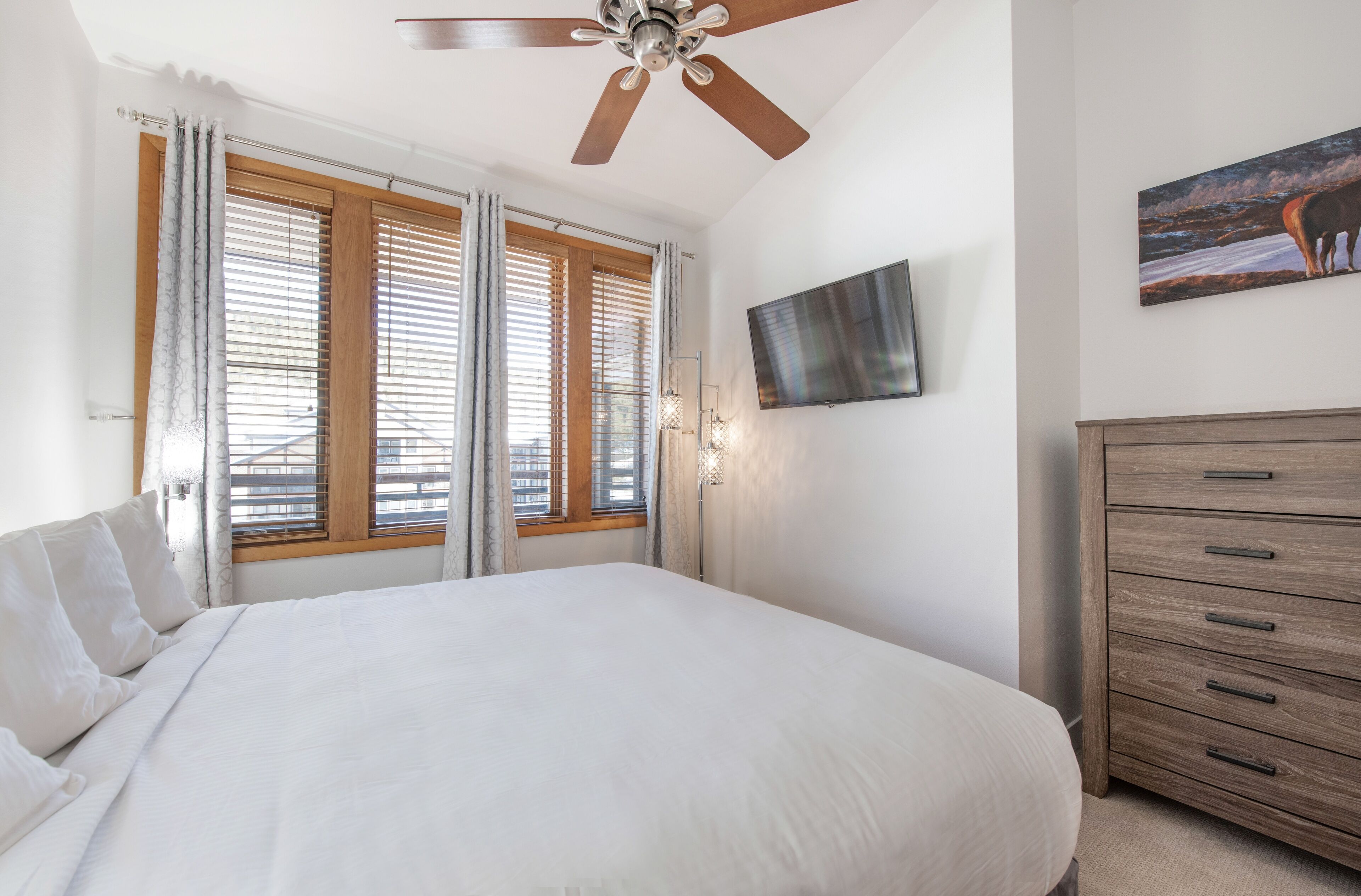 A bedroom with a large bed, a ceiling fan, a TV mounted on the wall, and a wooden dresser.
