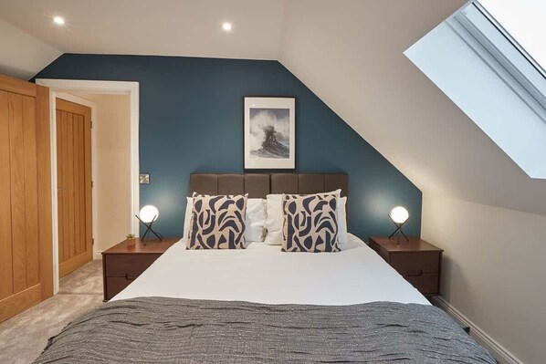 A bedroom with a sloped ceiling, a bed with patterned pillows, bedside lamps, and a framed picture on the wall. - Host & Stay | Iburn House (Briggswath)