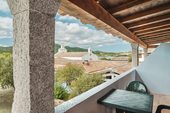 A balcony with a view of a town, featuring a stone pillar, a wooden ceiling, and a green plastic chair.