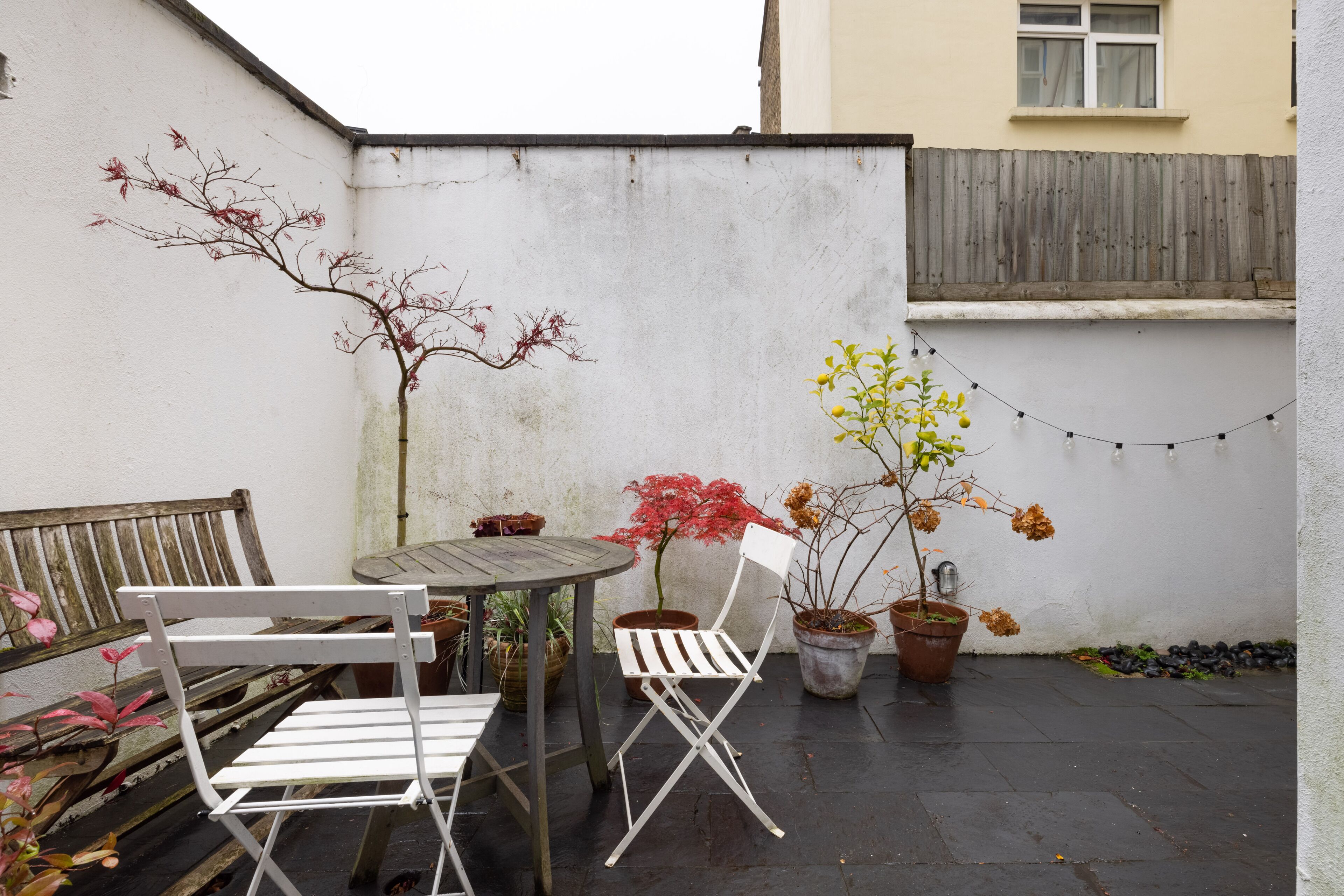 A small patio with a wooden table and two white chairs, surrounded by potted plants and a string of lights.