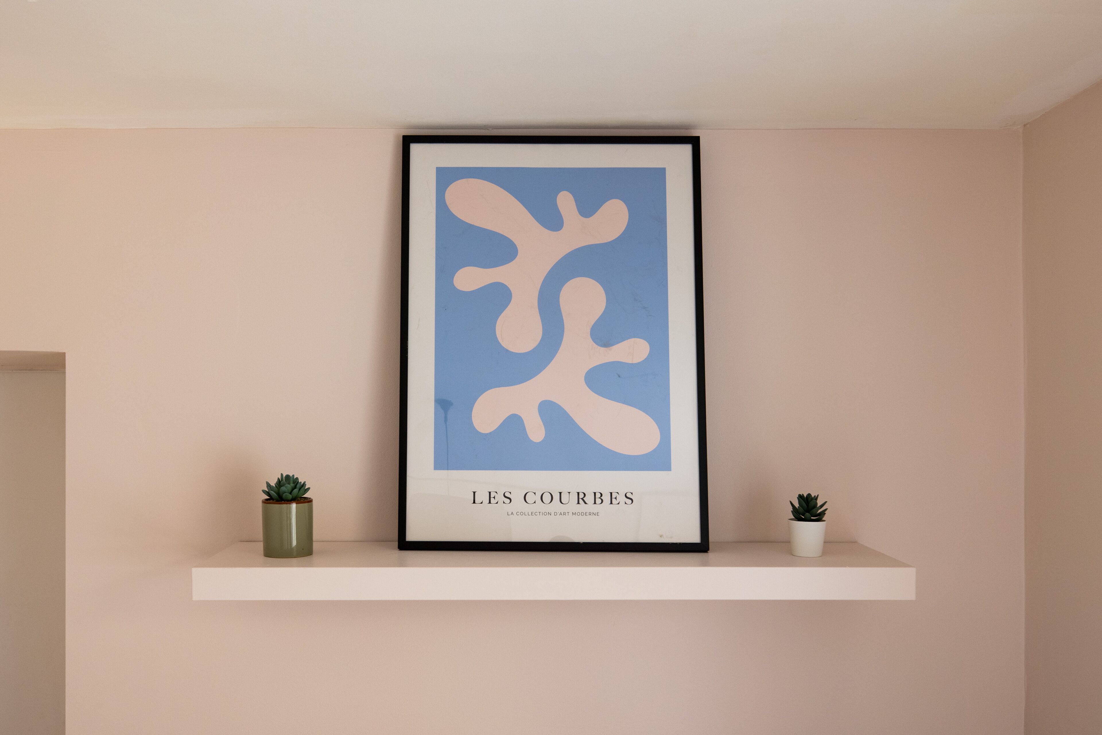 A framed artwork titled "LES COURBES" on a white shelf with two potted plants.