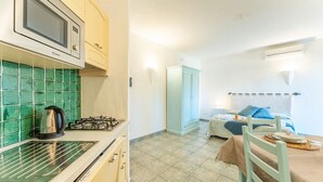A compact kitchen with a microwave, kettle, and gas stove. A dining area with a table and chairs is visible, and a bed is in the background. - Studio apartment 2 beds, about 30 square meters, living room with kitchenette and single beds that can be joined together, bathroom with shower. Possibility of cot (with supplement). <br>Comfortably furnished apartments, they can be on the ground floor w (Palau)