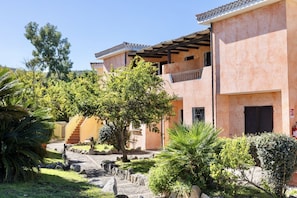 A building with a terracotta facade, a covered outdoor area with a staircase, and a variety of greenery including trees and shrubs.