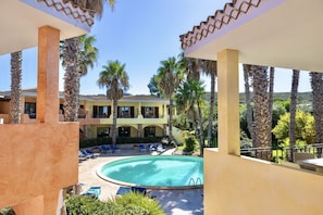 Zona de piscina con sillas de descanso y una alberca, rodeada de palmeras y un edificio con techo de tejas.