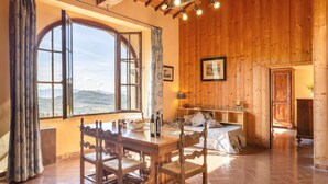 Ein gemütlicher Raum mit Holzboden, einem großen Fenster mit Vorhängen, einem Holzesstisch mit Stühlen und einem Bett mit Holzseitenverkleidung.