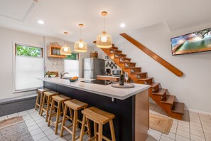 Een moderne keuken met een centraal eiland, houten krukken, hanglampen, een ingebouwde kookplaat en een houten trap.