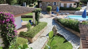 Una villa con un camino de piedra, una piscina y un jardín con una pérgola y sombrillas azules.