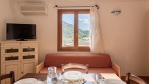Un área de comedor con una mesa lista para dos personas, un televisor sobre un mueble, y un ventanal con vista a un paisaje verde.