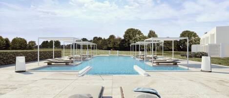 Un área de piscina con sillones de descanso y sombrillas, rodeada de árboles y un cielo despejado.