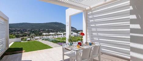 Un patio al aire libre con mesa y sillas, vistas a un paisaje verde y colinas distantes, y una estructura de pérgola blanca.