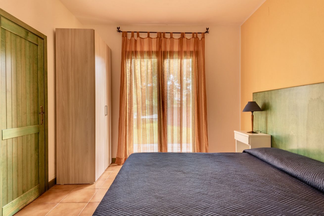 A bedroom with a bed, a nightstand with a lamp, a wooden wardrobe, and a door.