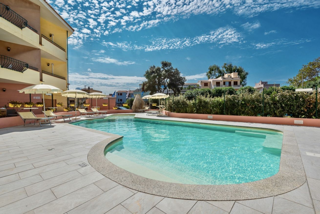 Ein Swimmingpool mit Liegestühlen und Sonnenschirmen, umgeben von einem Gebäude mit Balkonen und wolkenlosem Himmel.