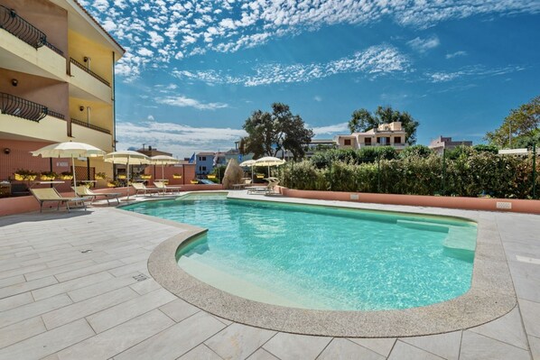 Une piscine entourée de chaises longues et de parasols, le tout bordé d’un bâtiment doté de balcons, sous un ciel dégagé.