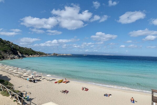 A beach with clear turquoise water, a sandy shore, and a rocky outcrop in the background.