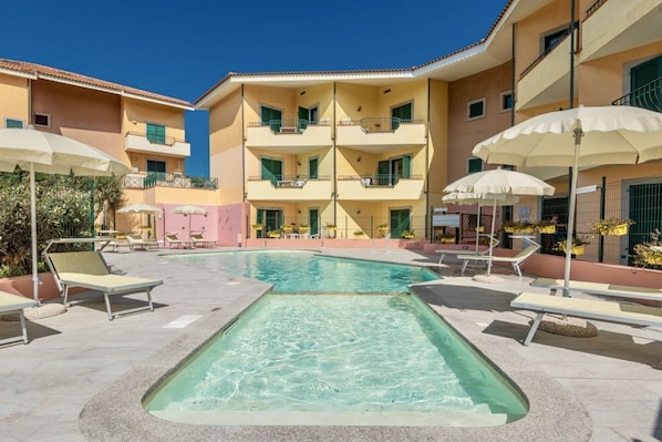 Un espace piscine doté de chaises longues et de parasols, entouré d’immeubles à plusieurs étages avec des balcons.