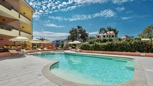 Une piscine entourée de chaises longues et de parasols, le tout bordé d’un bâtiment doté de balcons, sous un ciel dégagé.