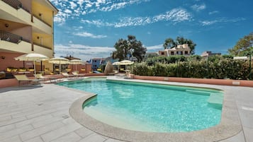A swimming pool with lounge chairs and umbrellas, surrounded by a building with balconies and a clear sky.