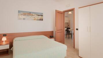 Una camera d'albergo con un letto, un comodino con una lampada, un quadro al muro e vista su un'area cucina attraverso una porta aperta.