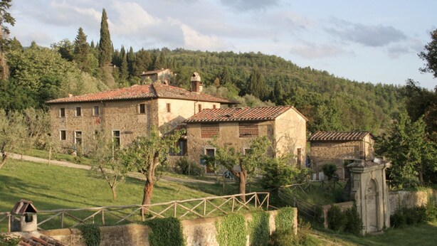 A rustic stone building with multiple windows and a tiled roof, surrounded by greenery and a stone wall.