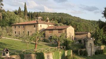 Un edificio rustico in pietra con numerose finestre e un tetto in tegole, immerso nel verde e circondato da un muro in pietra.