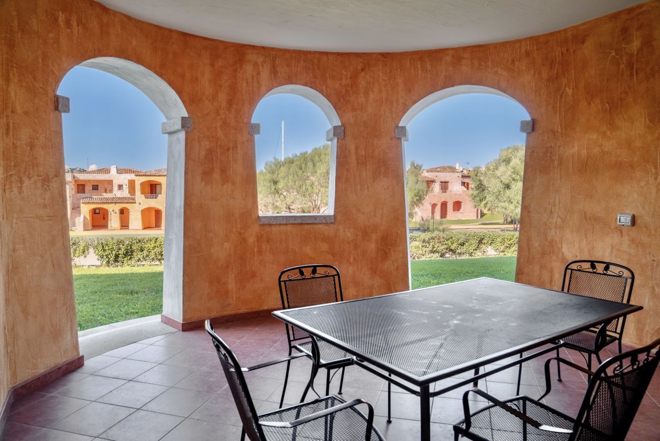 Un patio cubierto con mesa y sillas, amplias ventanas en arco y vistas a edificios y vegetación.
