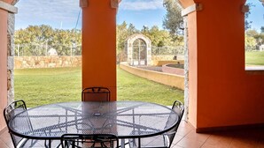 Une terrasse couverte avec une table et des chaises rondes en métal, des piliers en pierre et une vue sur un jardin clos.
