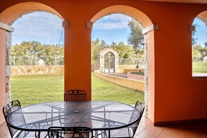 Un porche cubierto con una mesa y sillas de metal redondas, pilares de piedra y vistas a un jardín cercado.