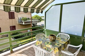 A balcony with a table and chairs, a large window, and a view of a building with balconies and greenery.