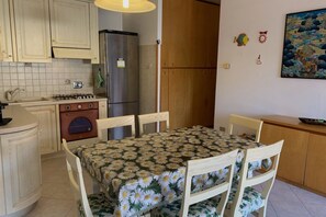 A compact kitchen with a refrigerator, stove, and wooden cabinets. There is a dining table with chairs, and a painting on the wall.