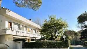 Um edifício de dois andares com uma varanda, uma árvore no jardim frontal e um céu azul límpido.