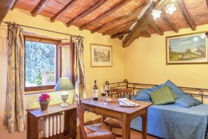 A cozy room with a wooden table, a bed with a blue cover, a window with curtains, a radiator, and a painting on the wall.