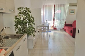 A compact kitchen with a sink, cabinets, and a dining area with a table and chairs. A potted plant is in the corner, and a red sofa is in the living area.