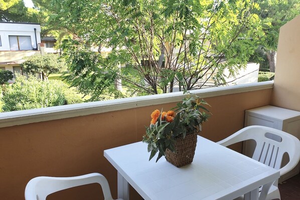 A balcony with a white table and chairs, a wicker basket with flowers, and a view of greenery and buildings.