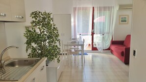 Een kleine keuken met een spoelbak, kastjes en een eethoek met een tafel en stoelen. In de hoek staat een potplant, en er is een rode bank in de woonkamer.