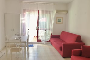 A room with a dining table, chairs, a red sofa, a white curtain, and a window with a view.