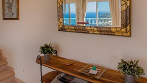 Ein Zimmer mit einem Holztisch, einer Topfpflanze, einem gerahmten Bild und einem großen Spiegel, der einen Balkon mit Meerblick reflektiert.