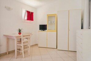 A small, clean room with a white table, a chair, a TV on a cabinet, and a window with red curtains.