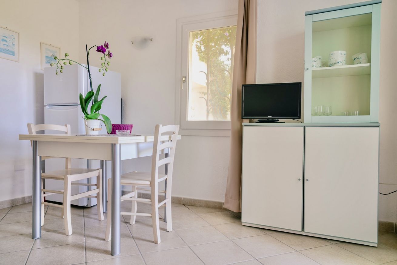 Une petite cuisine avec une table blanche, des chaises, un réfrigérateur, un téléviseur posé sur un meuble et une fenêtre avec des rideaux.