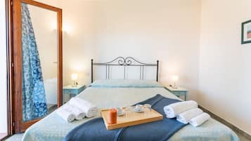 Una cama bien hecha con una colcha azul estampada, una bandeja de madera con toallas y una vela, y dos lámparas de noche.