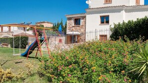 Eine weiße Villa mit steinverkleideter Fassade, ein umzäunter Garten mit bunten Blumen und ein Spielplatz mit einer Rutsche.